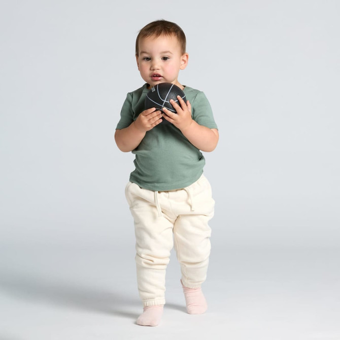 A toddler wears a green t-shirt and cream-coloured pants, holding a small ball in front of a light background.