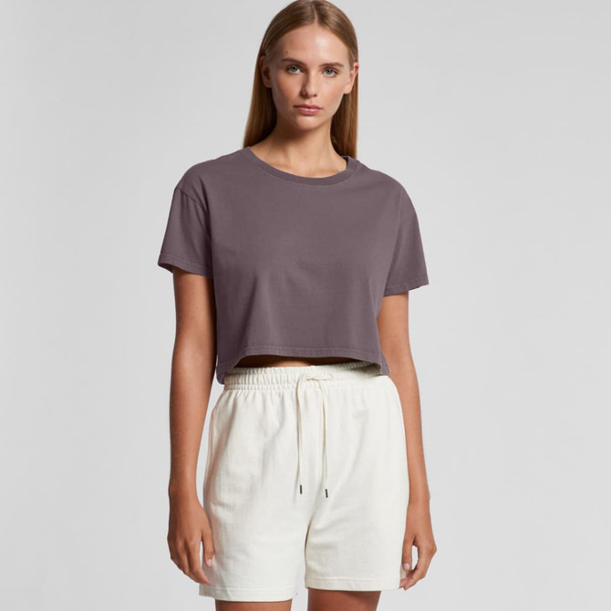 A model wears a faded crop tee in a muted purple colour, paired with light-coloured shorts. The shirt has a logo.
