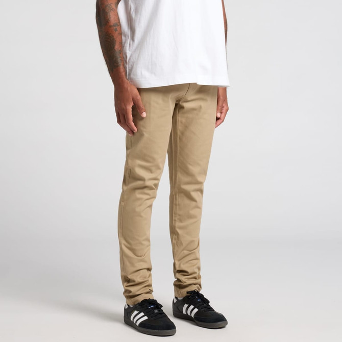 A pair of beige standard pants displayed on a person wearing a white shirt and black shoes, featuring a logo.