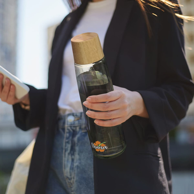 A drink bottle featuring a green glass body and a wooden cap, held by a person outdoors. The bottle has a logo.
