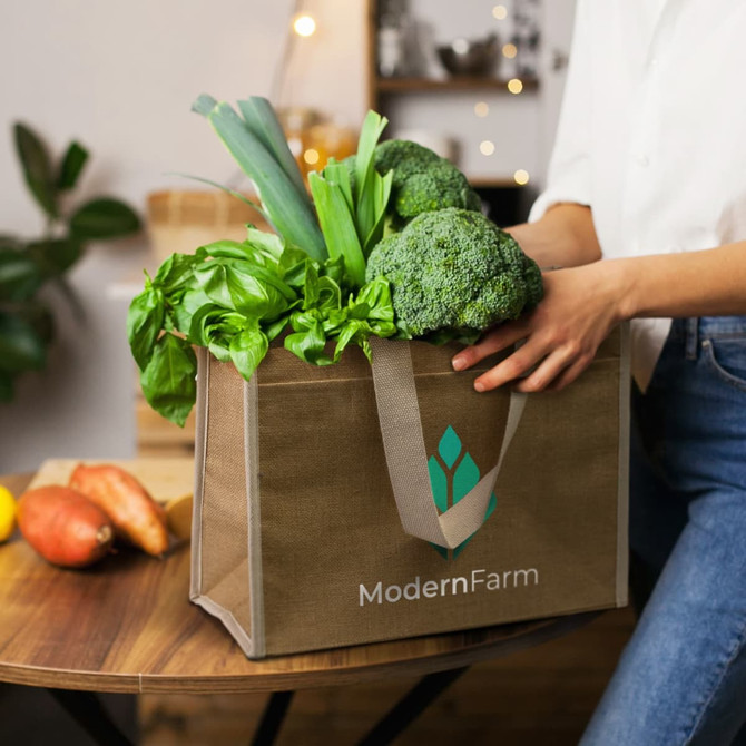 A jute cooler bag in brown, filled with fresh vegetables like broccoli and herbs, featuring a logo on the front.