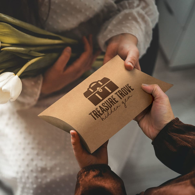 A large pillow box in brown cardboard, featuring a logo design, held by two hands with a bouquet of tulips.