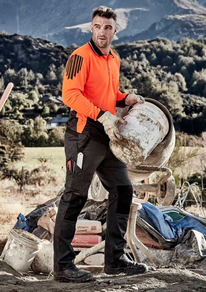 A man in an orange shirt and black tough work pants carries a bucket at a construction site, with mountains in the background.