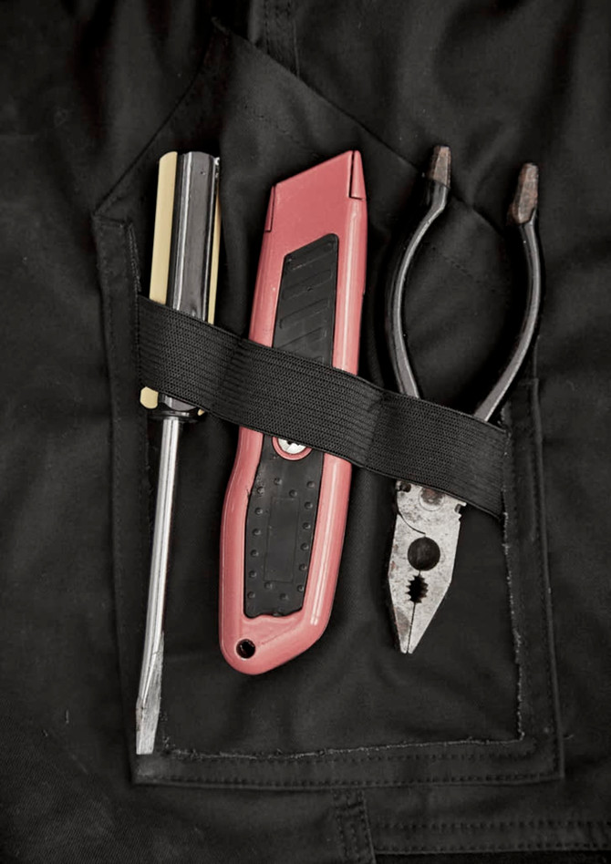 A close-up of a black work pant pocket featuring a screwdriver, utility knife, and pliers secured with a strap.