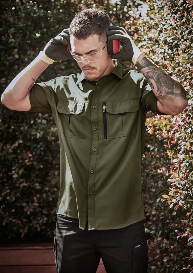 A man in an olive green short sleeve button-up shirt, wearing gloves and protective ear muffs, stands outdoors.