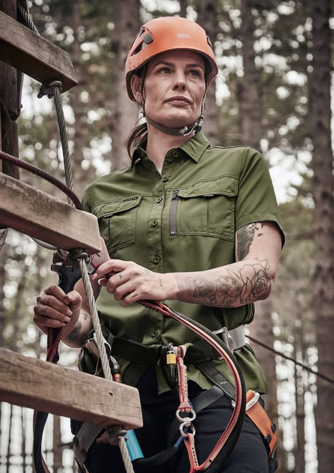 A woman in an olive green short sleeve shirt stands near a climbing structure, wearing a helmet and harness.