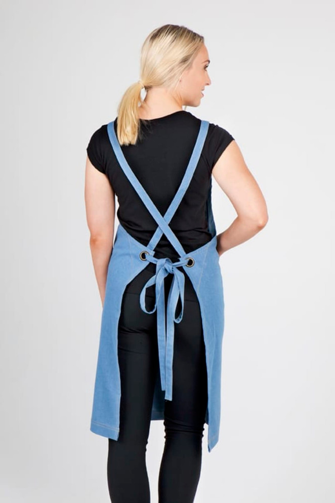 A full body denim apron in blue, featuring adjustable straps and a tied waist, displayed on a model from the back.