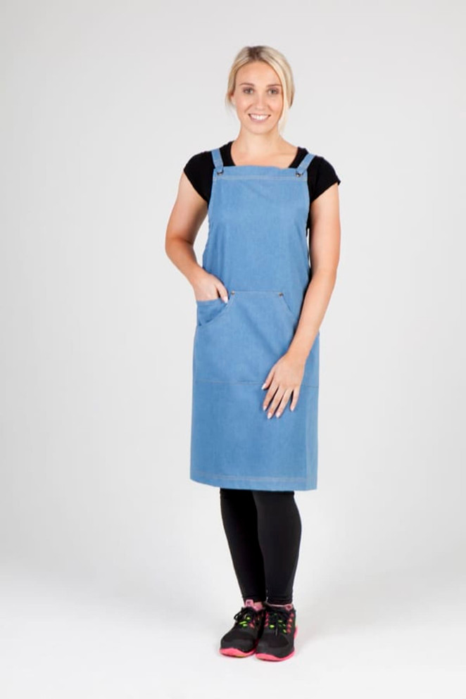 A woman wears a full body denim apron in blue, featuring straps and a front pocket, with black leggings and shoes.