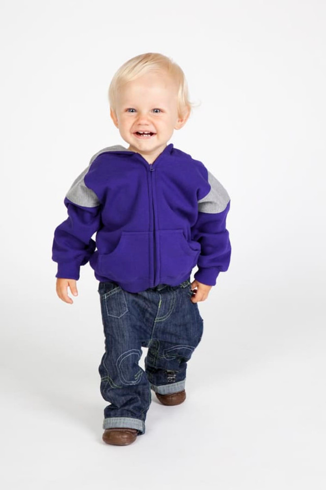 A smiling toddler in a purple hoodie with grey panels and jeans, walking against a plain white background.