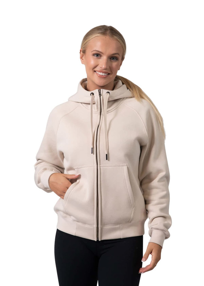 A women's cotton zippered hoodie in beige with a kangaroo pocket and a logo. Model is smiling and wearing black leggings.