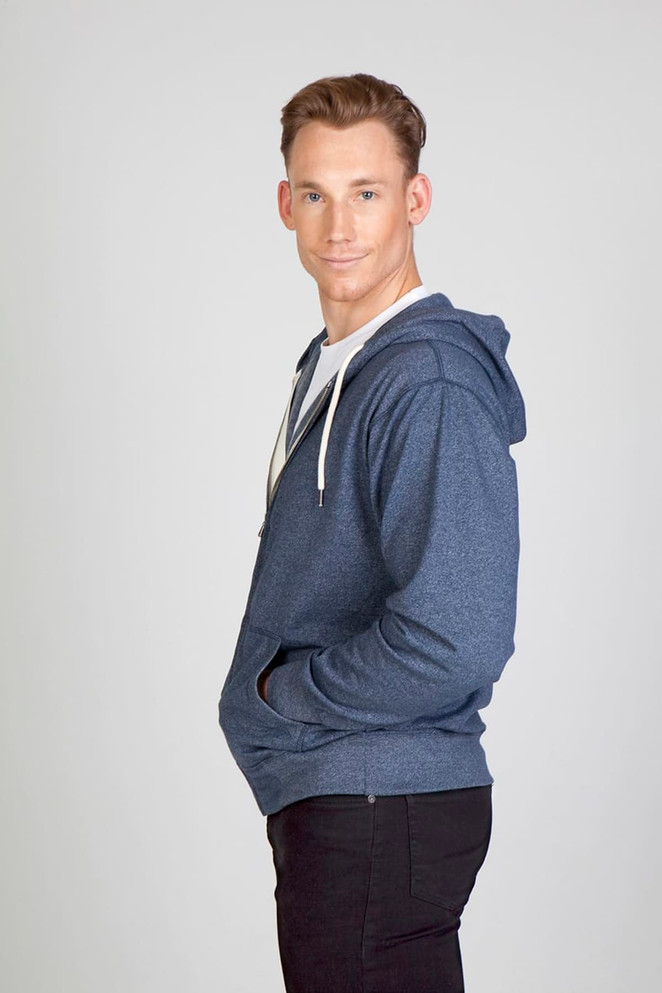 A man wearing a navy blue heather zip hoodie, standing with his hands in the pockets and smiling.