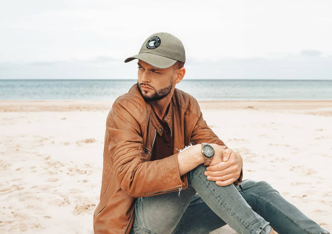 A man with a cap sits on the beach, wearing a brown jacket and grey jeans. The cap is olive green with a logo.