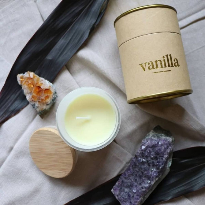 A medium candle in a tan container with a gold lid, surrounded by crystals and dark leaves, featuring a vanilla scent.