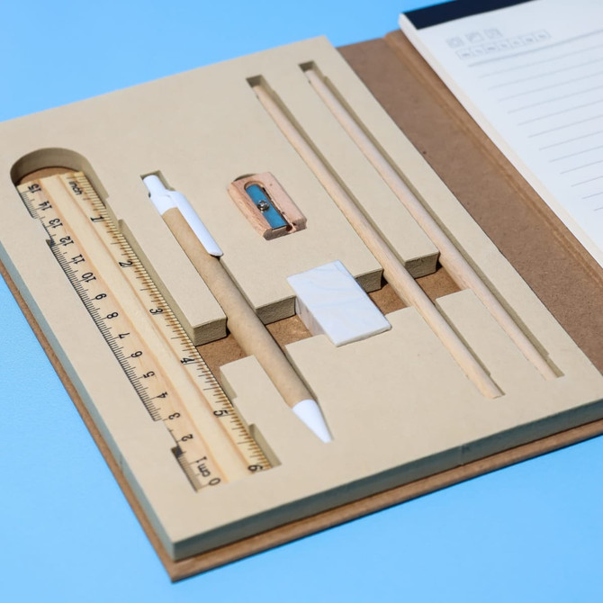 An A5 notepad in a brown cover with a wooden ruler, pencil, eraser, and sharpener, all arranged neatly inside.
