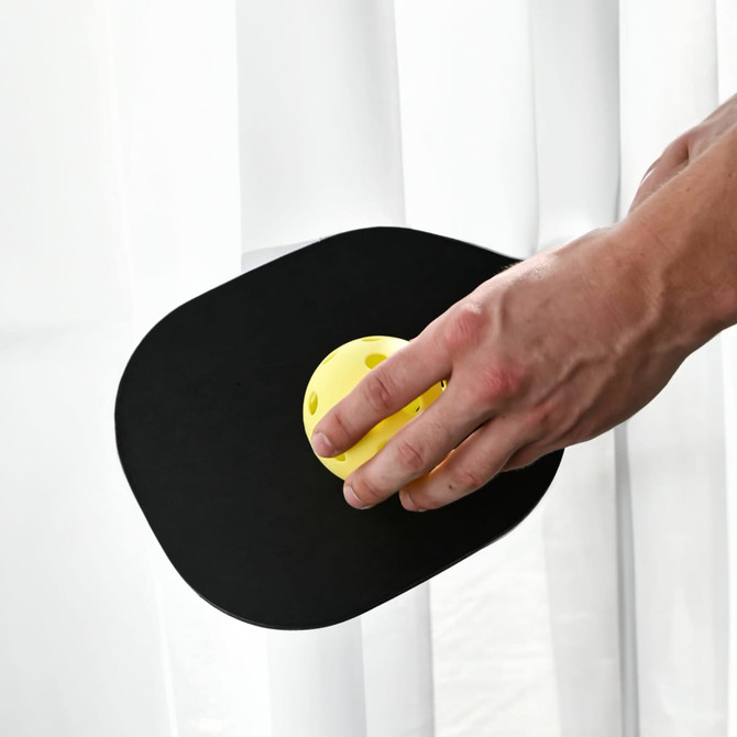 A paddle with a black surface and a yellow pickleball rests against a white curtain.