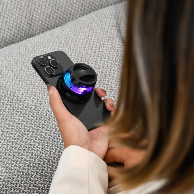 A compact, black speaker with colourful lights rests on a smartphone in a person's hand. The background features a textured fabric.