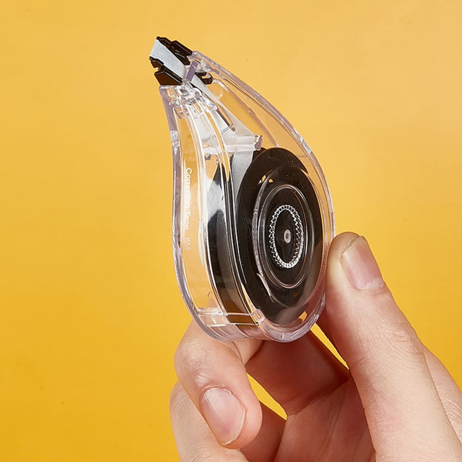 A hand holds a transparent correction tape dispenser with a black tape reel against an orange background.