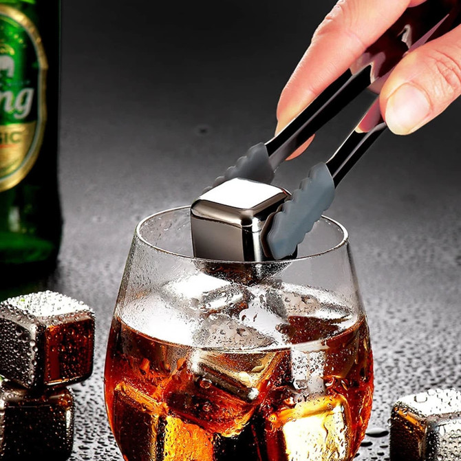 A drink glass filled with dark liquid and ice cubes, with a person's hand using tongs to place a metal ice cube.