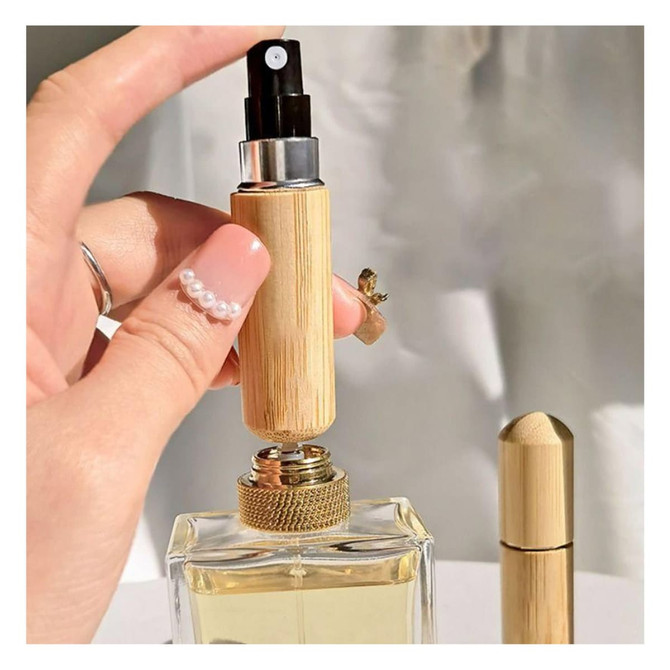 A mini bamboo atomiser held in a hand, with a perfume bottle in the background. The product features a natural bamboo finish.