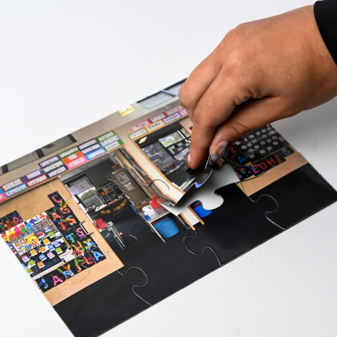 A hand places a piece into an A5 magnetic jigsaw featuring a colourful indoor scene with various items.