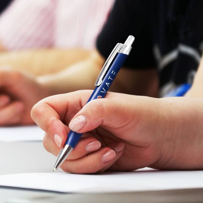 A metal pen in blue held by a hand, with a notepad and other people visible in the background.