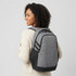 A grey laptop backpack with black accents, worn by a person with curly hair, featuring adjustable straps and side pockets.