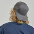A grey trucker cap with a mesh back, viewed from behind, showcasing curly hair and a navy shirt.