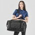 A woman holds a black duffle bag with a shoulder strap, featuring a logo and multiple compartments.