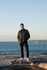A man stands by the ocean wearing a black puffer jacket, blue jeans, and white shoes, with a coastal background.