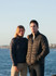A couple stands by the water wearing navy puffer jackets. The scene is coastal with a clear sky.