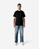 A man stands wearing a black Gildan Light Cotton T-Shirt and light blue jeans, with white shoes on a plain background.