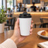 A reusable white coffee cup with a black lid, held in a hand, set against a café background.