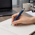 A blue metal pen is being held above a notebook with handwritten notes, alongside a laptop and coffee mug.