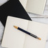 A metal pen with a blue barrel and wooden accents resting on an open notebook with blank pages.