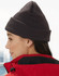 Acrylic beanie in dark grey worn by a person with long hair, paired with a red jacket. The beanie has a logo.