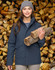 A woman in a navy quilted waterproof jacket holds logs, standing in front of a stacked woodpile. The jacket features a hood.