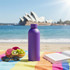 A drink bottle in purple stands on a beach towel, with the Sydney Opera House and a fruit bowl in the background.