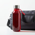 A drink bottle in a vibrant red colour with a silver lid and a handle, positioned next to a grey bag.