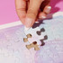 A hand placing a white jigsaw puzzle piece into a pastel-coloured puzzle on a pink background.