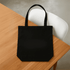 A black tote bag with two handles, placed on a wooden table. The bag has a logo on the front.