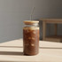 A glass tumbler with a bamboo lid and straw, featuring dark beverage and ice cubes, set on a wooden table.