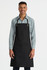 A black bib apron with adjustable straps, featuring a front pocket, worn by a model in a checkered shirt.