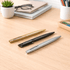Three metal ballpoint pens in gold, black, and silver arranged on a wooden desk with stationery in the background.