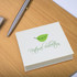 A square sticky note pad in cream with a green leaf design, resting on a wooden surface beside a pen.
