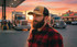 A man with a beard wears a tan and black trucker cap, standing beside trucks at a gas station during sunset.
