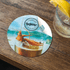 A round coaster featuring a vibrant illustration of prawns on a drink, with a glossy finish and a logo.