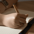 A hand holding a pen writes in a lined, cream-coloured notebook on a wooden surface.