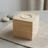 A square wooden candle holder with a light-coloured wood finish and a small white candle on top, placed on a textured surface.