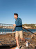 A man in a blue long-sleeve shirt and shorts walks near a rocky shoreline, with a waterfront city backdrop.