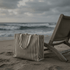 A striped tote bag in beige and cream rests on the sand beside a wooden beach chair, with waves in the background.